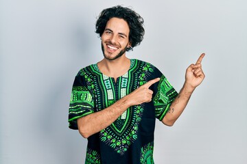 Poster - Handsome hispanic man wearing traditional african clothes smiling and looking at the camera pointing with two hands and fingers to the side.