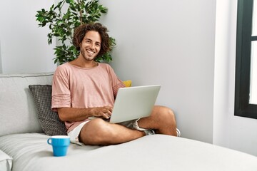 Canvas Print - Young hispanic man smiling happy using laptop sitting on the sofa at home.