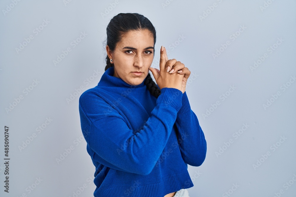 Young hispanic woman standing over isolated background holding symbolic gun with hand gesture, playing killing shooting weapons, angry face