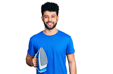 Poster - Young arab man with beard holding electric steam iron looking positive and happy standing and smiling with a confident smile showing teeth