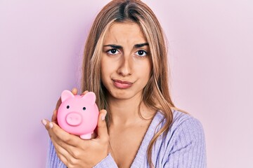 Sticker - Beautiful hispanic woman holding piggy bank skeptic and nervous, frowning upset because of problem. negative person.