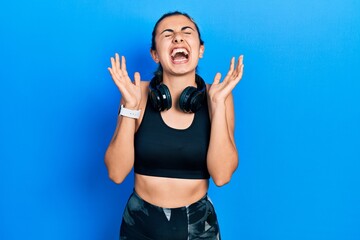 Poster - Beautiful hispanic woman wearing gym clothes and using headphones celebrating mad and crazy for success with arms raised and closed eyes screaming excited. winner concept