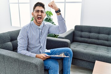 Sticker - Young psychologist man at consultation office angry and mad raising fist frustrated and furious while shouting with anger. rage and aggressive concept.