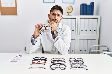 Sticker - Young optician man holding optometry glasses looking confident at the camera smiling with crossed arms and hand raised on chin. thinking positive.