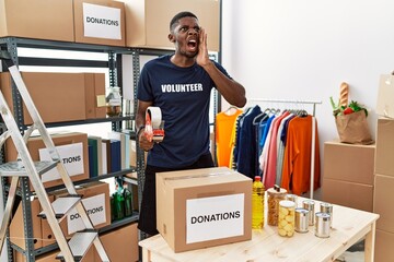 Poster - Young african american volunteer man packing donations box for charity shouting and screaming loud to side with hand on mouth. communication concept.