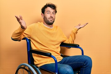 Poster - Handsome man with beard sitting on wheelchair clueless and confused expression with arms and hands raised. doubt concept.