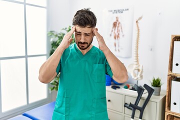 Poster - Young man with beard working at pain recovery clinic with hand on head for pain in head because stress. suffering migraine.