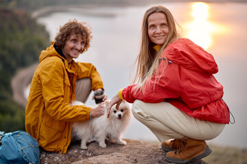 couple with pet dog Hiking in mountains together. Adventure. river in the background. Excited man and woman drinking tea during hiking, travelling, spending time together outdoors in nature