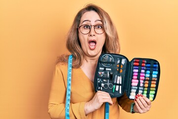 Poster - Middle age caucasian woman wearing tape measure holding sew kit afraid and shocked with surprise and amazed expression, fear and excited face.