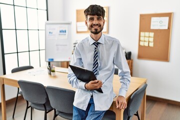 Wall Mural - Hispanic man with beard wearing business style sitting on desk at office looking positive and happy standing and smiling with a confident smile showing teeth