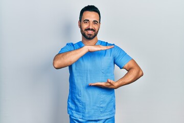 Poster - Handsome hispanic man with beard wearing blue male nurse uniform gesturing with hands showing big and large size sign, measure symbol. smiling looking at the camera. measuring concept.