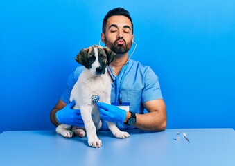 Wall Mural - Handsome hispanic veterinary man with beard checking dog health using stethoscope looking at the camera blowing a kiss being lovely and sexy. love expression.