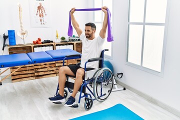 Wall Mural - Young hispanic man patient having rehab session using elastic band sitting on wheelchair at clinic