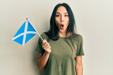 Wall Mural - Young hispanic girl holding scotland flag scared and amazed with open mouth for surprise, disbelief face