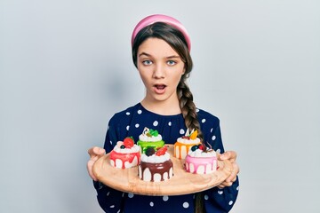 Poster - Young brunette girl holding sweet pastries in shock face, looking skeptical and sarcastic, surprised with open mouth