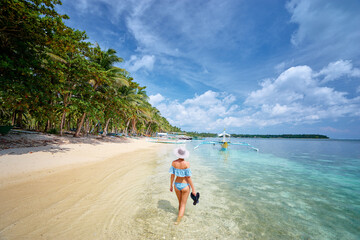 Wall Mural - Vacation on the seashore. Back view of young woman walking away on the beautiful tropical white sand beach.