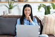 © kucherav - Young smiling Asian woman sitting on sofa using laptop to browse social networks, shopping. Having a good time at home