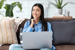 © kucherav - Young smiling Asian woman sitting on sofa using laptop to browse social networks, shopping. Having a good time at home