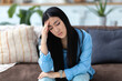 © kucherav - Asian young woman sitting on the sofa at home holding her head, headache, migraine, emotional stress