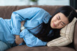 Asian young woman suffering from abdominal pain while lying down on sofa at home