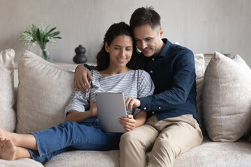 Wall Mural - Smiling young couple using tablet together, hugging relaxing sitting on couch, happy woman and man looking at smartphone screen, shopping or chatting online, spending leisure time with gadget at home