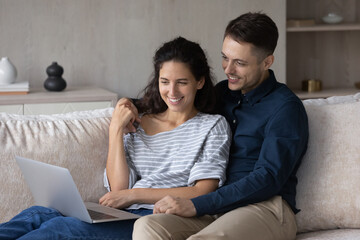 Wall Mural - Happy couple using laptop together, relaxing on couch at home, smiling young woman and man holding hands, looking at computer screen, watching movie or funny video, spending leisure time with gadget