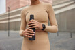 Cropped shot of unrecognizable woman dressed in sportswear holds sporty bottle of fresh water wears smartwatch feels thirsty after cardio training stands outdoors against blurred background.