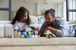 © fizkes - Interested small cute curly African American kid girl constructing building with wooden blocks with caring young father, sitting together on floor carpet in living room, enjoying playing at home.