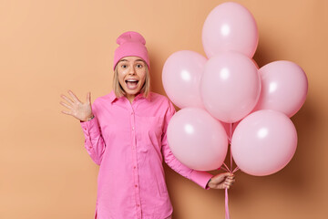 Canvas Print - Indoor shot of emotional woman wears pink hat and shirt exclaims loudly holds bunch of helium balloons comes on birthday party isolated over beige background. Festive event and celebration concept