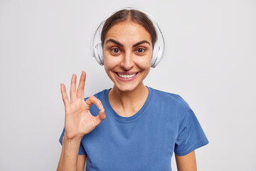 Wall Mural - Attractive brunette woman makes okay gesture says I agree smiles happily wears stereo headphones and casual blue t shirt isolated over white background. Teenage girl assures everything is good