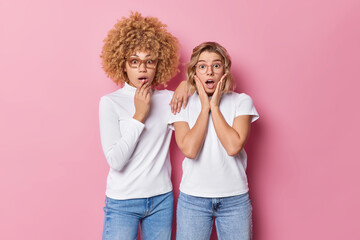 Wall Mural - Indoor shot of two scared young women stare emotive feel shocked wear casual clothes cannot believe own eyes react on something amazing stare a camera isolated over pink background. Omg concept
