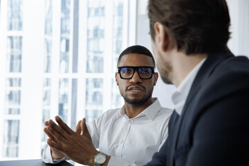 Sticker - Serious engaged African employee in glasses talking to coworker, business partner, speaking, telling about project. Multiethnic workers, professionals discussing work tasks at workplace