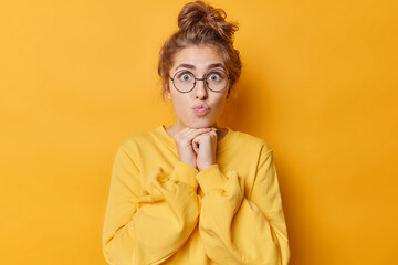 Poster - Portrait of surprised lovely woman keeps lips rounded hands under chin has dark hair gathered in bun wears round spectacles and jumper isolated over yellow background. Human face expressions