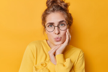 Poster - Portrait of lovely tender young woman has folded lips keeps hand on cheek looks gently at camera wears round transparent glasses and jumper poses against yellow background has romantic expression