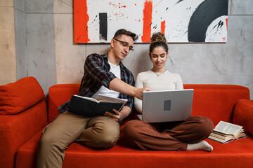 Man and woman young adult couple generation Z sitting together on the sofa bed at home study male and female friends helping each other with lesson tutor and student learning education concept