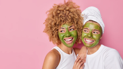 Horizontal shot of happy women apply green nourishing masks on face look gladfully away stand closely to each other isolated over pink background blank copy space for your advertising content