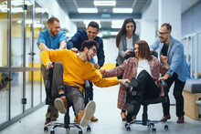 Young Cheerful Business People  Having Fun While Racing On Office Chairs And Smiling. Friendly Work Team Enjoying Fun Work Break Activities.