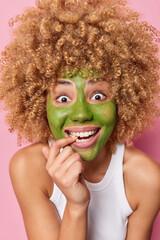 Canvas Print - Vertical shot of happy young curly blonde woman applies green cucumber mask smiles broadly bites finger wears casual t shirt poses indoor. Beauty facial treatments and cosmetic procedures concept