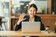 © PaeGAG - Smiling asian businesswoman waving hand doing video chat looking at laptop consulting customer sitting at workplace
