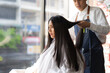 © Siam Stock - Yong long-haired Asian female client in white haircut cape sitting on hairdressing chair looking at herself in the mirror while getting a haircut by young Asian male hairdresser in hair salon.
