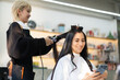 © Siam Stock - Smiling cute blond Asian female hairdresser in denim apron straightening hair of Caucasian female client in white haircut cape sitting on hairdressing chair smiling using a cellphone in the hair salon