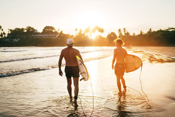 Wall Mural - Father with teenager son walking with surfboards by the sandy ocean beach with palm trees on background lightened with sunset sun. They smiling and have a conversation. Family active vacation concept.