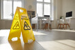 © Studio Romantic - Close up of yellow standing plastic sign with human figure that slips and falls and words Caution Wet Floor placed by janitorial service in empty clean sunny office with desks, chairs and computers