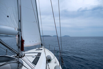 Wall Mural - Sailing in Aegean sea, Greece. Sailboat with open white sail, cloudy sky and rippled sea.