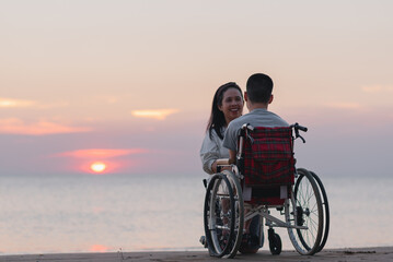 Behind of young man with disability and parent or volunteer or caregiver looking sunset on the sea beach at sunset with travel in summer, Positive photos give life energy and power concept.