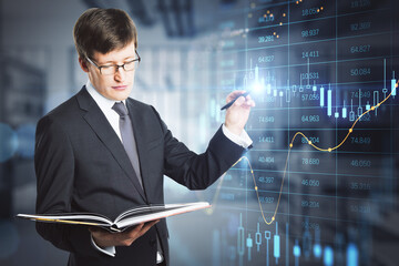 Poster - Portrait of attractive young businessman with accounting book pointing at abstract glowing big data forex candlestick chart on blurry office interior background. 