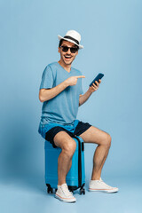 Excited charismatic guy in sunglasses and a hat, sits on a blue suitcase with a smartphone in his hand, male tourist prepares for a trip, looks at the camera with happy smile. Time for vacation