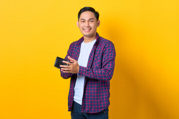 Wall Mural - Smiling young Asian man in plaid shirt holding wallet isolated on yellow background