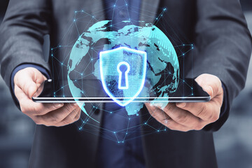 Close up of male hands holding tablet device with abstract glowing globe hologram with security shield icon on blurry office interior background. 