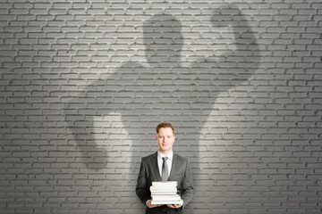 Canvas Print - Abstract image of attractive european businessman with books and strong muscle shadow on brick wall background. Confidence and strength concept.